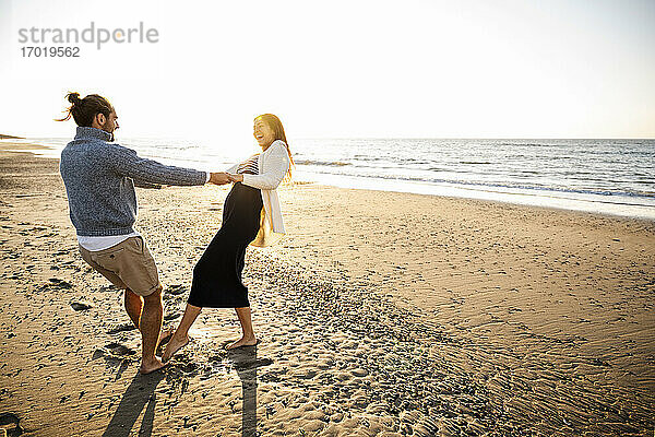 Fröhliches Paar hält sich an den Händen und tanzt am Strand gegen den klaren Himmel an einem sonnigen Tag
