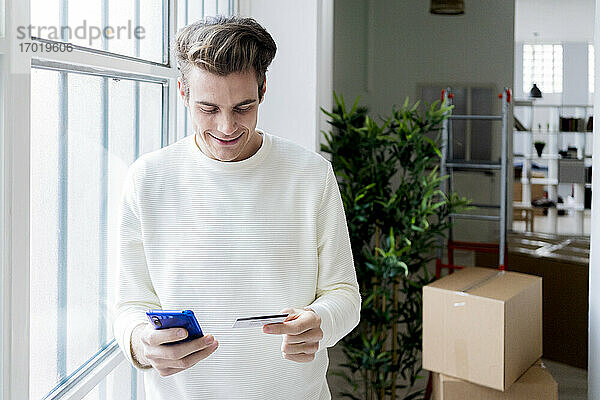 Lächelnder Mann  der in seiner neuen Loft-Wohnung mit Kreditkarte und Smartphone bezahlt