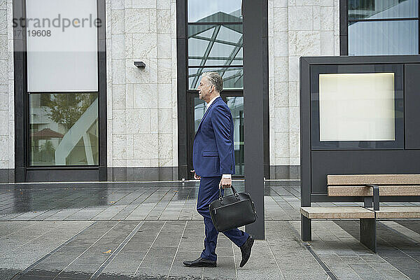 Älterer männlicher Unternehmer  der eine Tasche hält  während er auf einem Fußweg in der Stadt spazieren geht