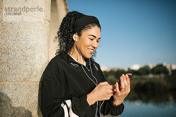 Lächelnde Sportlerin mit Handy  die Musik hört  während sie sich an eine Säule lehnt  an einem sonnigen Tag