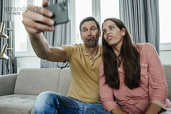 Glückliches Paar macht Gesichter bei der Aufnahme eines Selfies  während es zu Hause auf dem Sofa sitzt