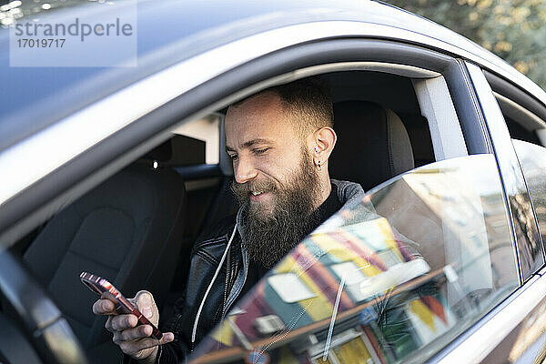 Bärtiger Mann benutzt Mobiltelefon  während er im Auto sitzt und durch das Fenster gesehen wird