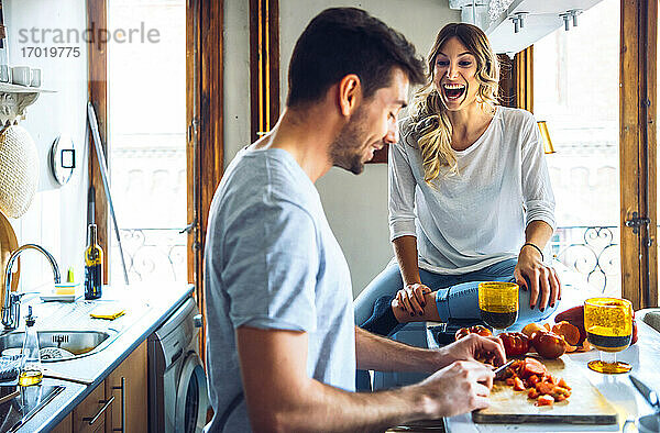Junger Mann bereitet Essen mit lachender Freundin in der Küche zu Hause zu