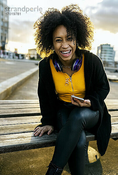 Fröhliche junge Frau mit Afro-Haar sitzt auf einer Bank in der Stadt bei Sonnenuntergang