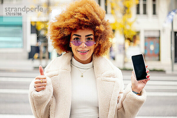 Lächelnde Frau mit Afro-Haar  die ein Smartphone in der Hand hält  während sie in der Stadt steht