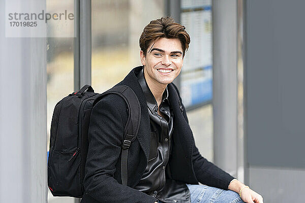 Lächelnder gut aussehender junger Mann  der mit seinem Rucksack an der Bushaltestelle sitzt und wegschaut