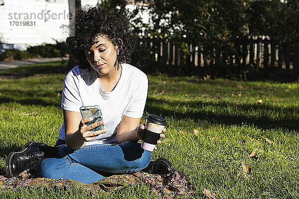 Junge Frau mit Einweg-Kaffeebecher und Mobiltelefon im Park sitzend