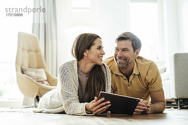 Lächelnder Mann und Frau mit digitalem Tablet  die sich gegenseitig ansehen  während sie zu Hause liegen