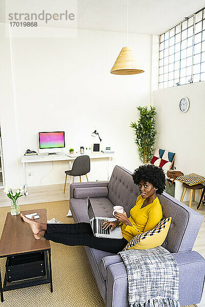 Junge Frau mit Laptop trinkt Kaffee  während sie zu Hause auf dem Sofa sitzt