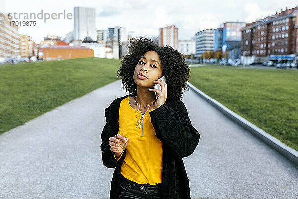 Junge Frau mit Afrohaar  die auf einem Fußweg in der Stadt steht und über ein Smartphone spricht