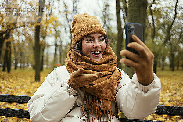 Glückliche junge Frau mit Strickmütze bei einem Videogespräch im Herbstpark