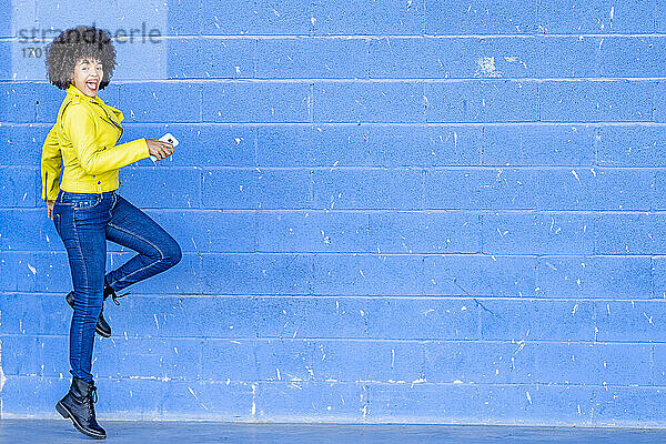 Fröhliche junge Frau mit Mobiltelefon tanzt gegen eine blaue Wand