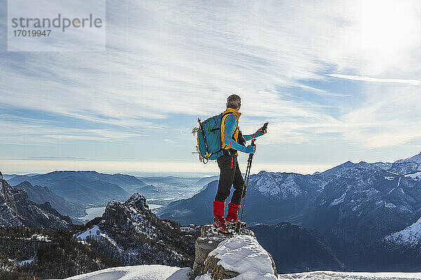 Männlicher Wanderer  der sein Smartphone benutzt  während er im Winter auf einem Berg gegen den Himmel steht  Orobische Alpen  Lecco  Italien