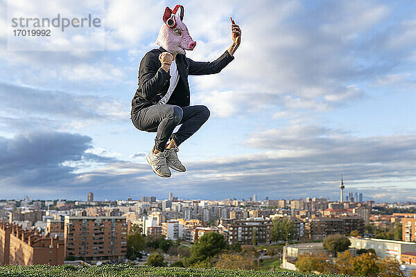 Männlicher Profi mit Schweinemaske  der ein Selfie mit seinem Smartphone macht  während er in der Stadt gegen den Himmel springt