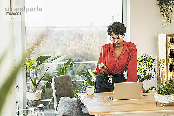 Frau schaut auf Smartphone am Tisch mit Laptop zu Hause