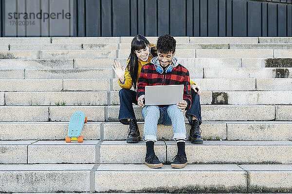 Lächelnde Frau mit winkender Hand bei einem Videogespräch über einen Laptop  während sie mit einem Freund auf einer Treppe sitzt