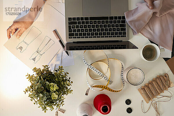Modedesignerin prüft Stoffmuster und entwirft am Laptop am Schreibtisch im Atelier