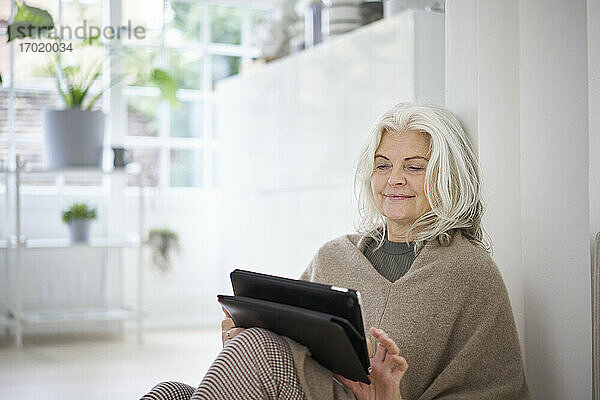 Lächelnde Frau mit weißen Haaren  die ein digitales Tablet benutzt  während sie im Wohnzimmer einer Wohnung sitzt