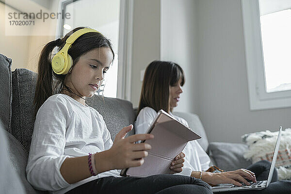 Tochter hält ein digitales Tablet  während die Mutter einen Laptop benutzt und zu Hause sitzt
