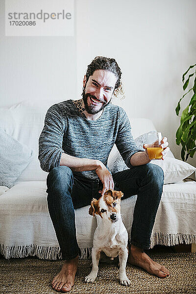 Lächelnder junger Mann hält ein Saftglas in der Hand und sitzt mit seinem Hund auf dem Sofa zu Hause
