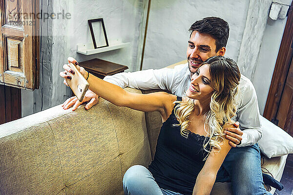 Lächelndes junges Paar nimmt Selfie auf Couch im Wohnzimmer zu Hause