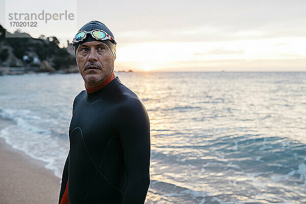 Porträt eines männlichen Schwimmers  der bei Sonnenuntergang allein am Küstenstrand steht