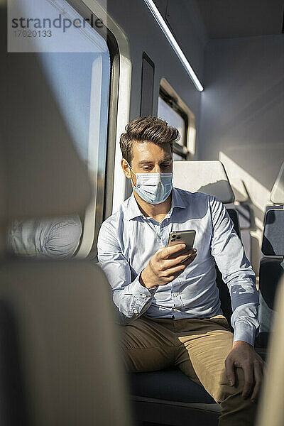 Geschäftsmann mit Schutzmaske  der im Zug sitzend ein Mobiltelefon benutzt  während COVID-19