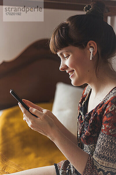 Lächelnde Frau  die einen Videoanruf auf einem Smartphone tätigt  während sie im Schlafzimmer zu Hause sitzt