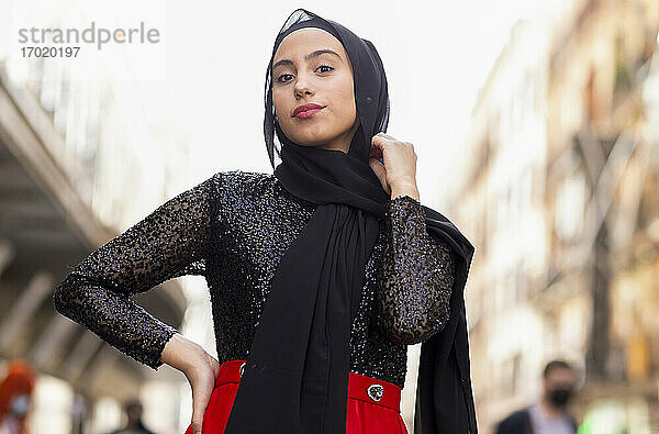 Porträt einer jungen Frau mit schwarzem Hidschab  die im Freien mit der Hand auf der Hüfte posiert