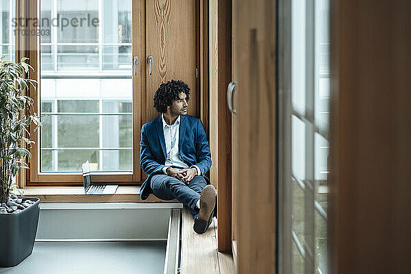 Nachdenklicher männlicher Unternehmer  der wegschaut  während er mit seinem Laptop vor dem Fenster im Büro sitzt