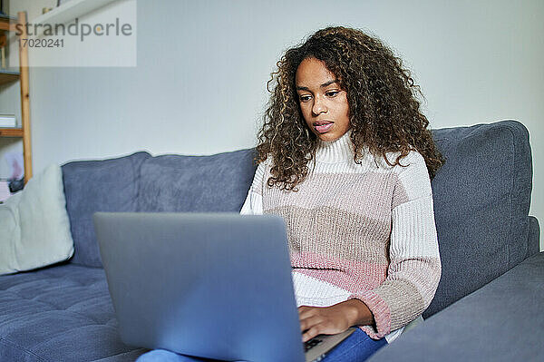 Junge afroamerikanische Frau  die auf dem Sofa zu Hause konzentriert einen Laptop benutzt