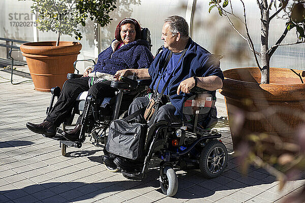 Behinderte Frau und Mann halten sich an den Händen  während sie an einem sonnigen Tag im Rollstuhl sitzen