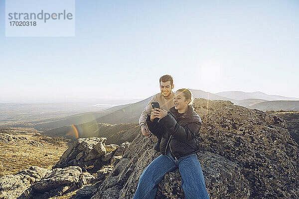 Freunde benutzen ihr Smartphone  während sie auf einem Berg gegen den klaren Himmel sitzen