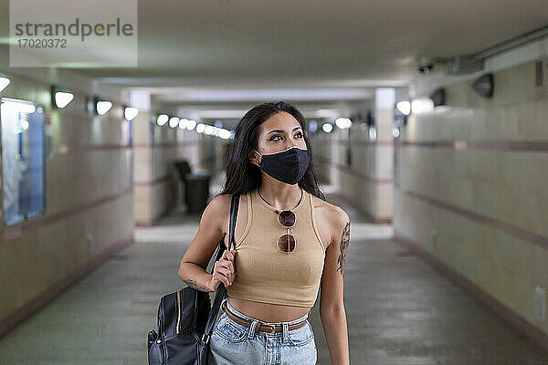 Junge Frau mit Gesichtsmaske  die eine Tasche trägt  während sie in einer unterirdischen Bahnhofspassage geht