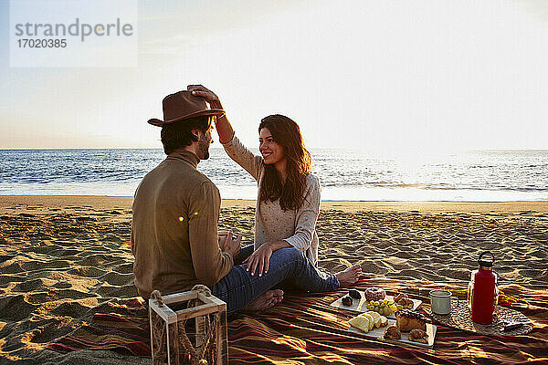 Frau spielt mit Hut und sitzt neben einem Mann am Strand