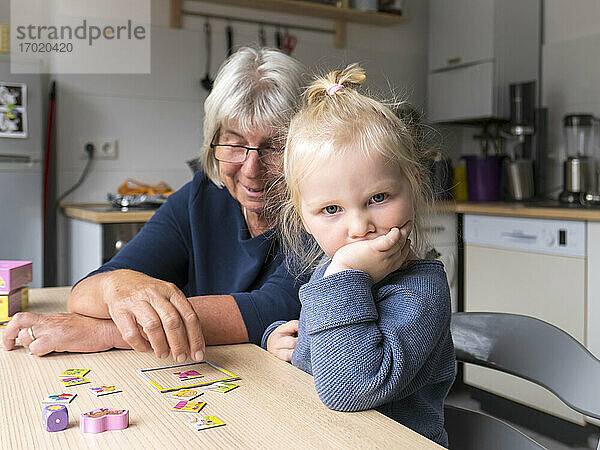Aufgeregtes blondes Mädchen sitzt bei der Großmutter und spielt Puzzle am Esstisch in der Küche