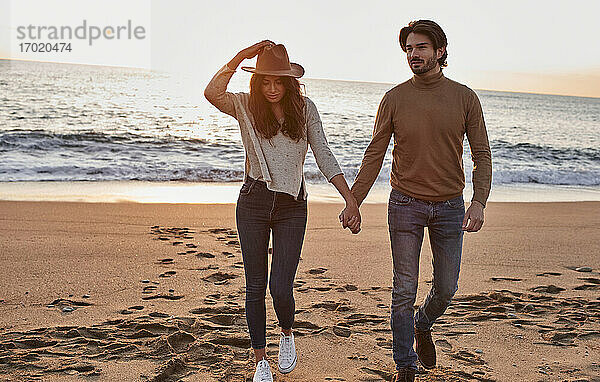 Junge Frau mit Hut hält die Hand eines Mannes  während sie gegen das Meer läuft