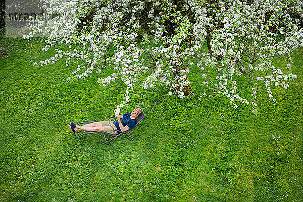 Ein Mann liest ein Buch auf einem Stuhl unter einem Baum im Garten