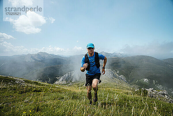 Älterer Sportler beim Trailrunning in den Bergen auf einer Wiese vor blauem Himmel