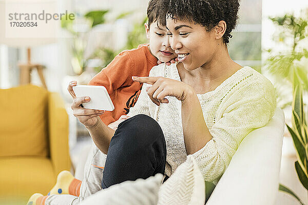 Lächelnde Mutter zeigt ihrem Sohn das Handy  während sie zu Hause sitzt