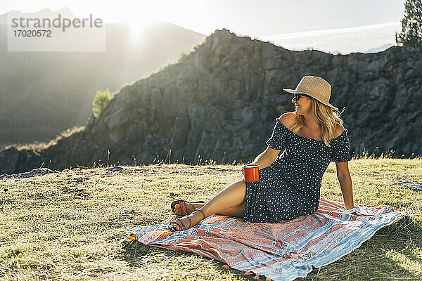 Lächelnde Frau mit Kaffeetasse träumt beim Sitzen auf einer Picknickdecke an einem sonnigen Tag