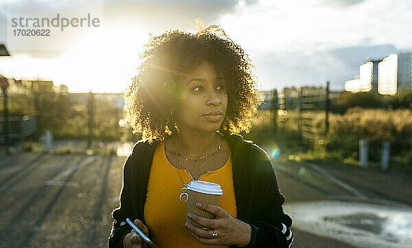 Junge Frau mit Afro-Haar hält Kaffee und Smartphone und schaut in den Himmel