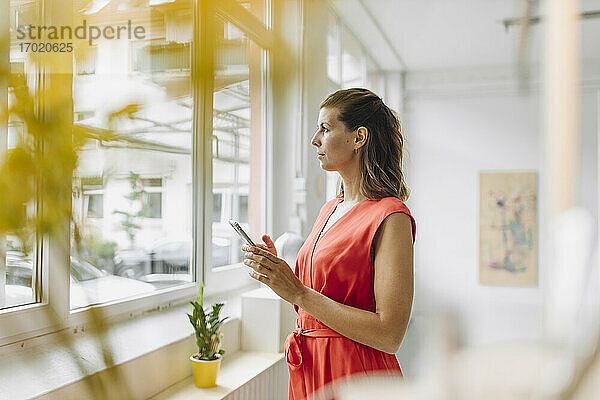 Geschäftsfrau hält digitales Tablet und schaut durch ein Fenster im Büro