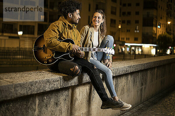 Paar mit Gitarre  das lächelnd auf einer Stützmauer sitzt