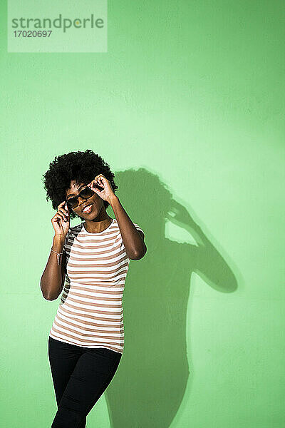Junge Frau passt ihre Sonnenbrille an  während sie vor einem grünen Hintergrund steht