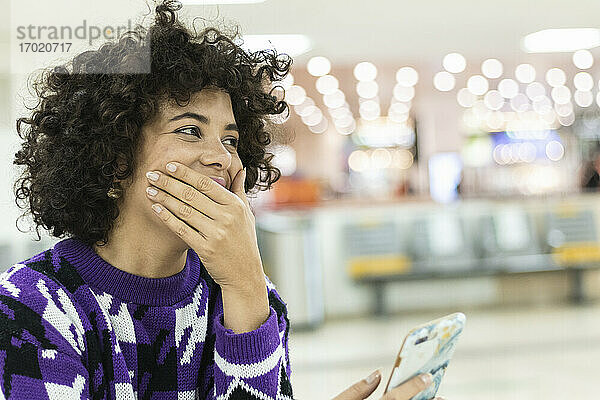 Junge Frau lacht  während sie am Flughafen sitzt und ein Mobiltelefon benutzt