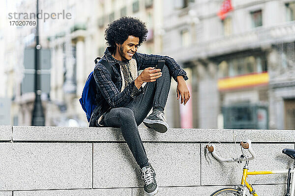 Lächelnder Mann mit lockigem Haar  der ein Mobiltelefon benutzt  während er auf einer Stützmauer in der Stadt sitzt