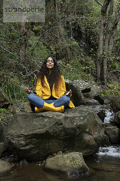 Wanderin in gelbem Regenmantel meditiert auf einem Felsen im Wald sitzend