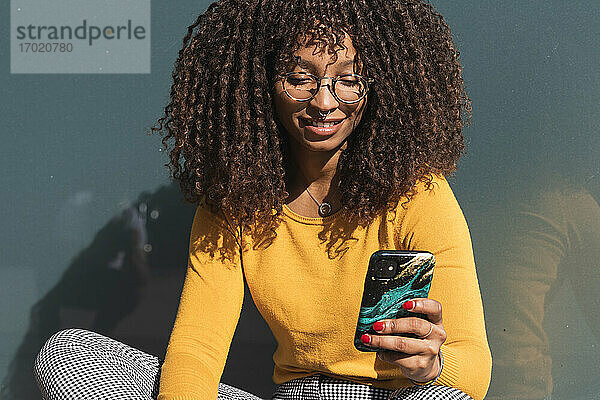 Lächelnde junge Frau  die ihr Mobiltelefon an einem sonnigen Tag gegen ein Glas hält