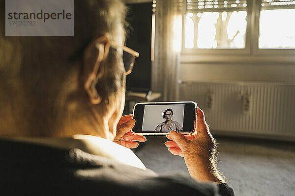 Ein älterer Mann lässt sich von einer Allgemeinmedizinerin per Videoanruf über ein Smartphone im Wohnzimmer beraten.